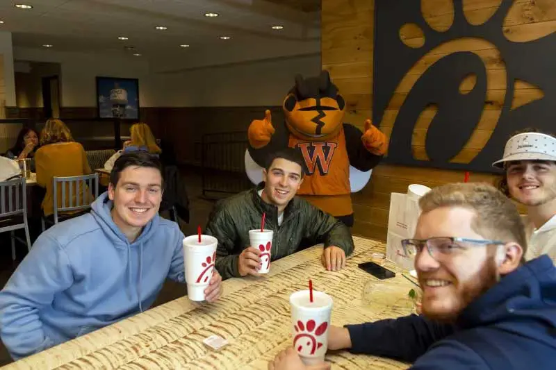 Students enjoying beverages at table inside Chick-fil-A with school mascot giving thumbs up in background